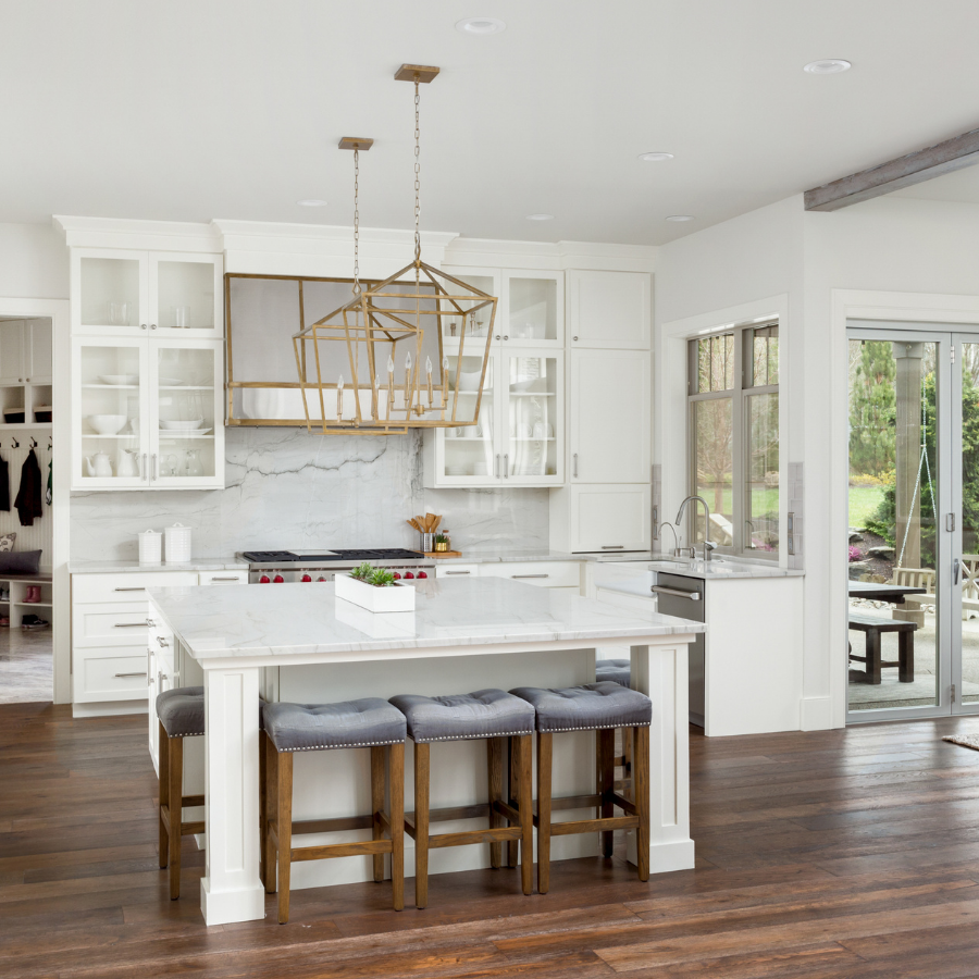 Bright white kitchen with a marble island and four wooden upholstered stools, a gold geometric chandelier, and sliding glass doors to a green yard.